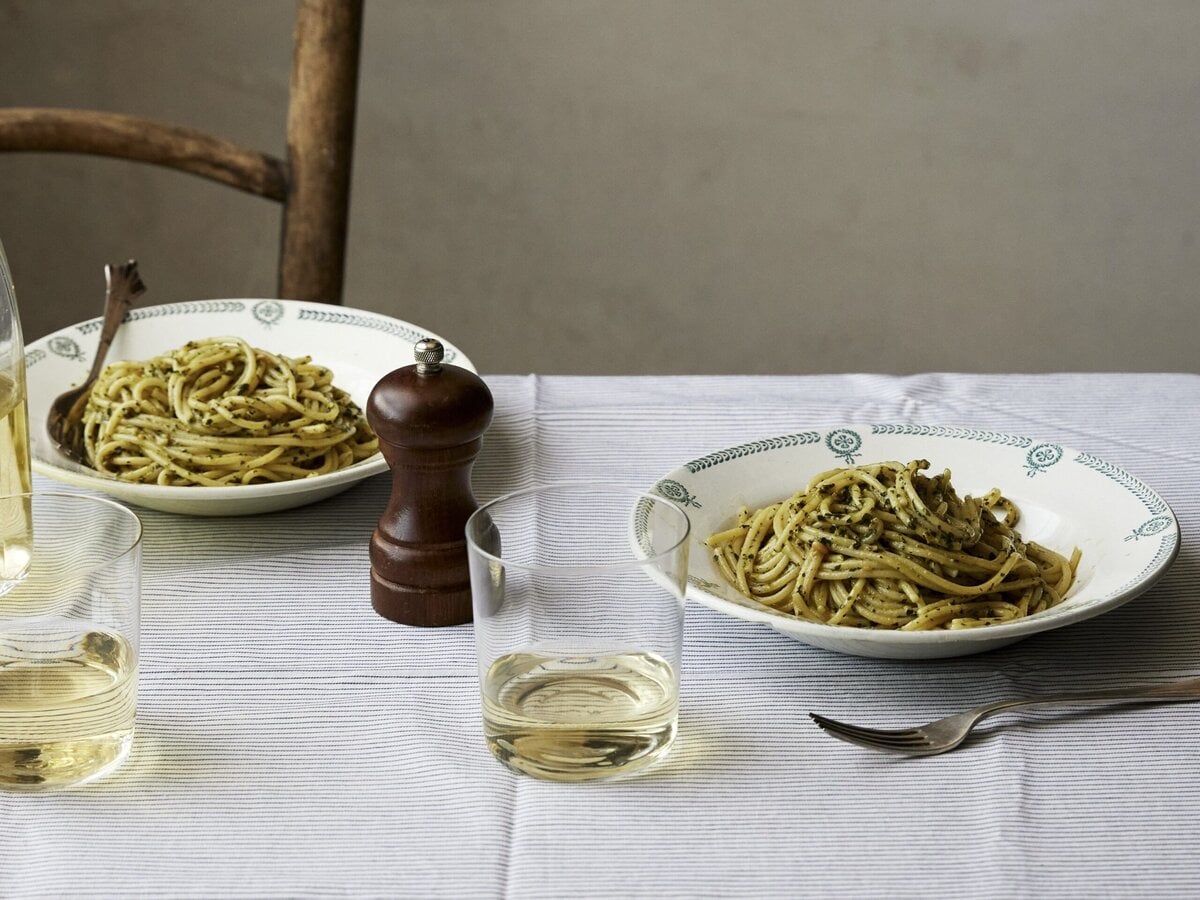 Gedekte tafel met twee borden spaghetti met pesto. Op tafel staat een pepermolen en glazen