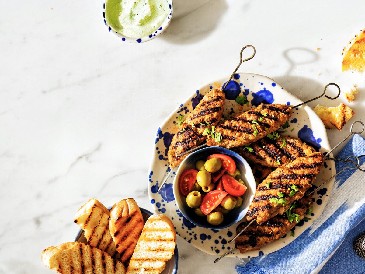 Köfte met bulgur aan een spies met een bakje olijven en tomaatjes, een bakje geroosterd brood en een bakje yoghurtknoflooksaus
