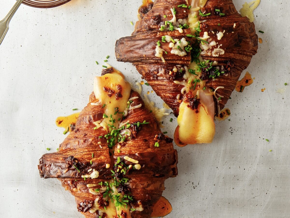 Croissants die in de lengte zijn doorgesneden met daartussen kaas en ham en daarover bieslook en crispy chili-olie