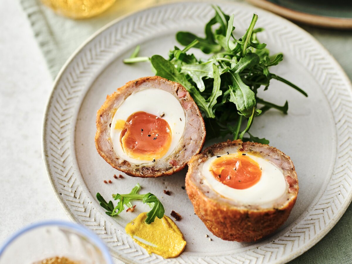 Een Italiaans gekruide scotch egg doormidden, met ei in het midden en daaromheen vlees, geserveerd met rucola en mosterd
