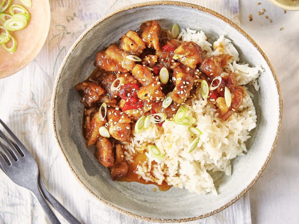 Twee borden rijst met kip met chilipeper en honing, met daarover lente-ui, rode peper en sesamzaad