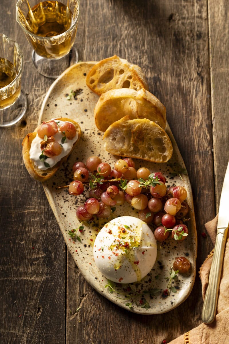 Bol burrata met olijfolie erover met daarnaast geroosterde rode druiven en sneetjes ciabatta