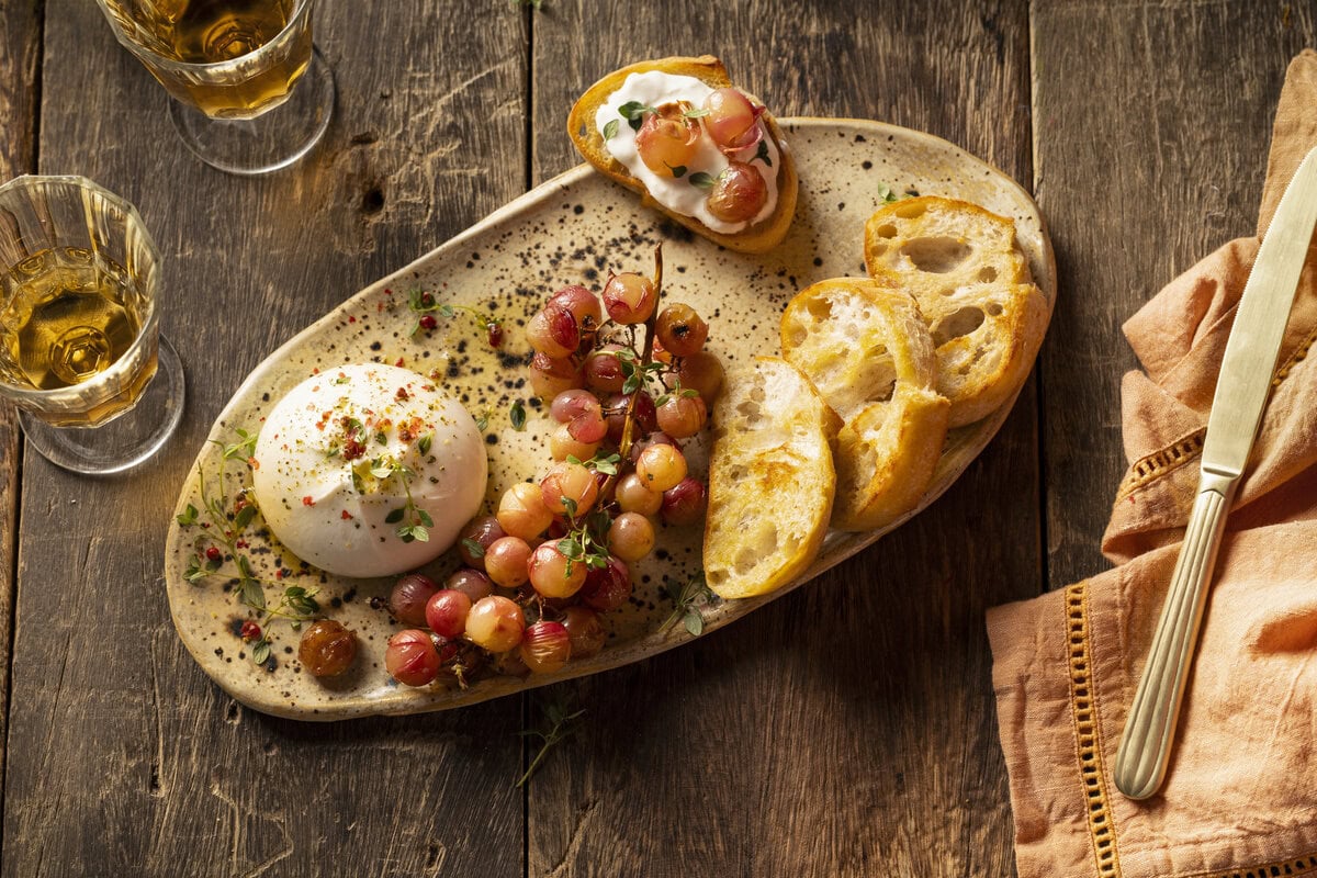 Bol burrata met olijfolie erover met daarnaast geroosterde rode druiven en sneetjes ciabatta